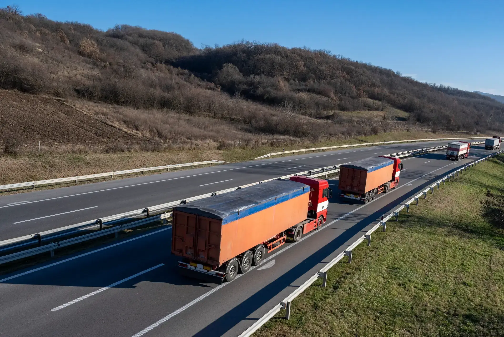Two orange trucks driving on a highway through a hilly landscape.