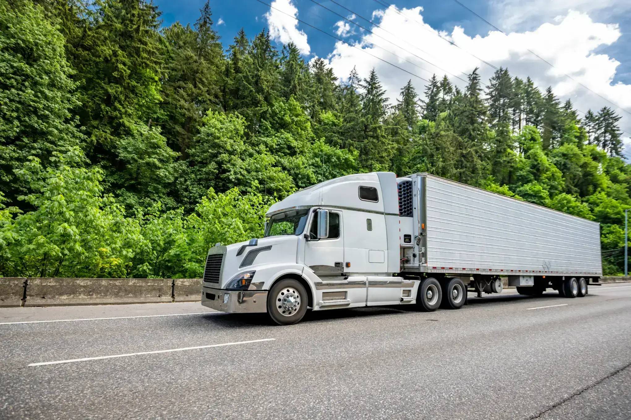 A white semi-truck driving on a highway surrounded by green trees.